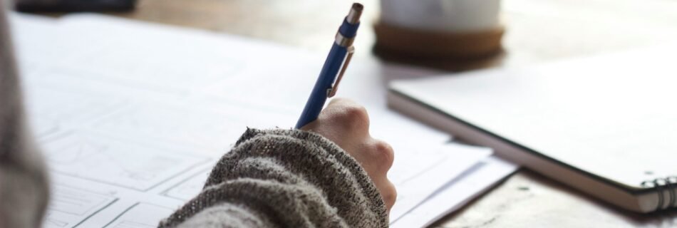 person writing on brown wooden table near white ceramic mug