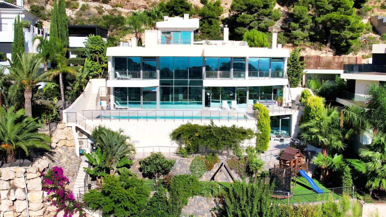 Luxury modern villa with glass facade, infinity pool, and lush garden on a hillside.